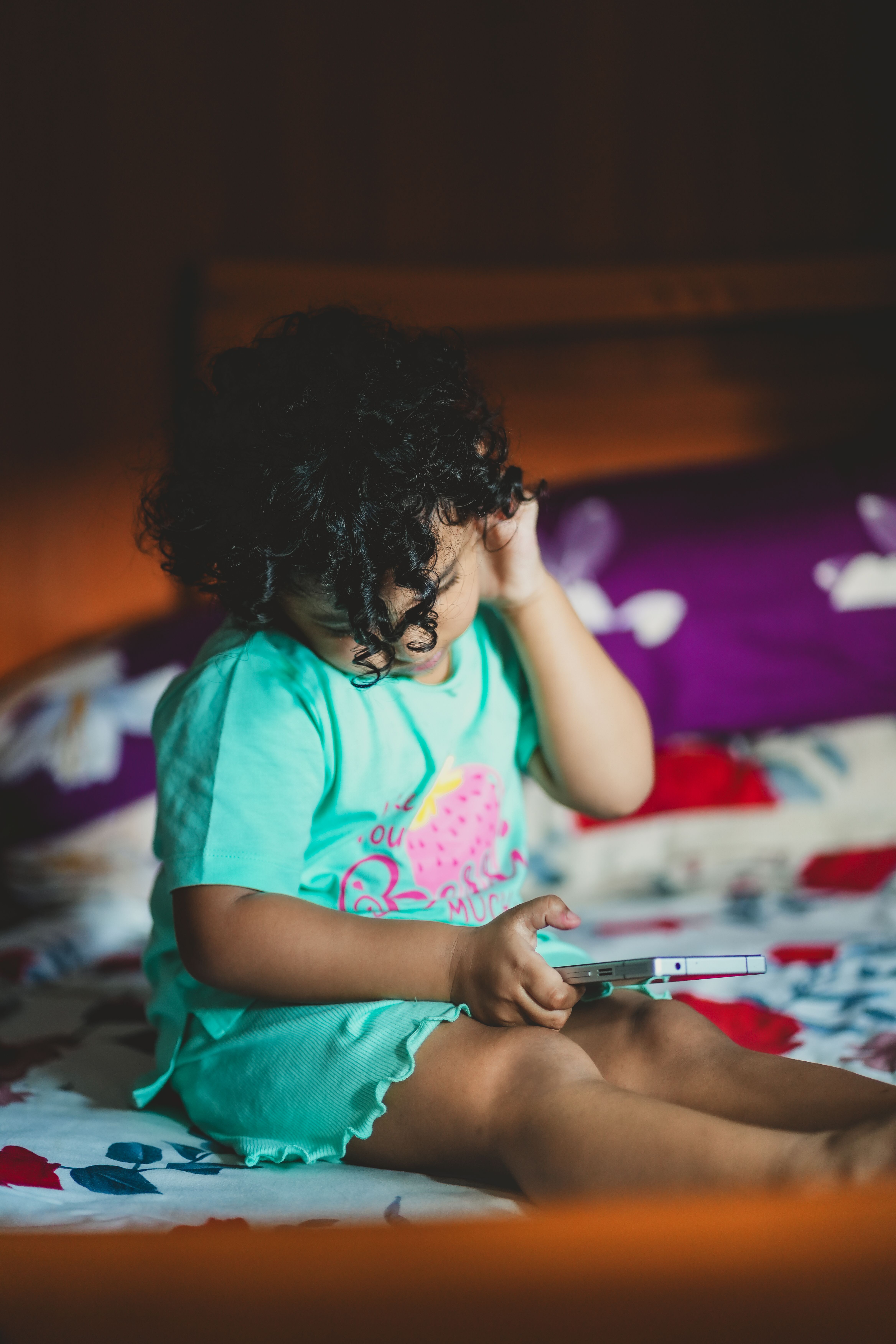 A young child in a teal outfit sits on a bed, holding a toy. The image captures a peaceful indoor moment.