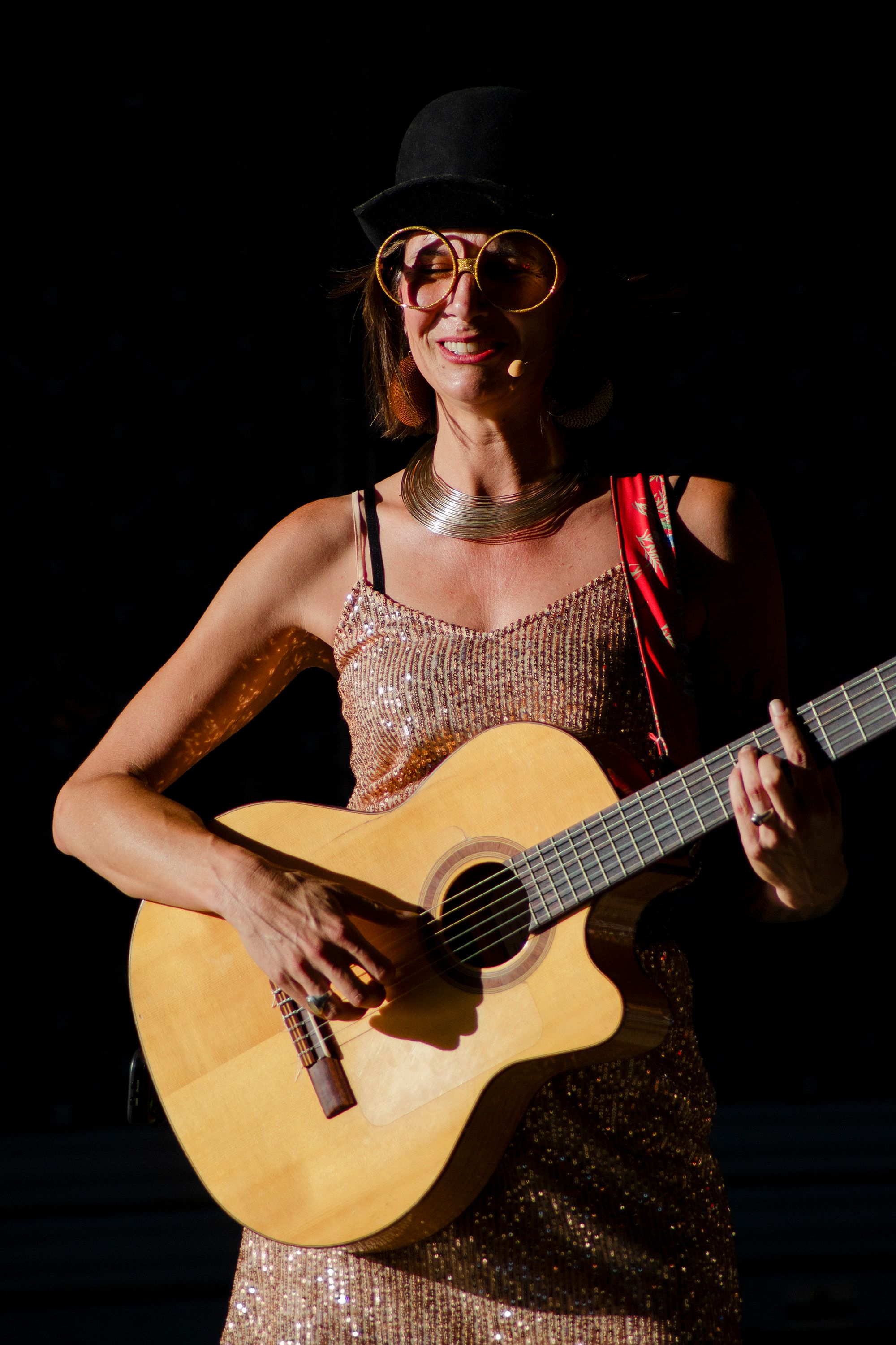 A cheerful woman plays guitar at an outdoor event in Périgueux, France, wearing a stylish hat and glasses.