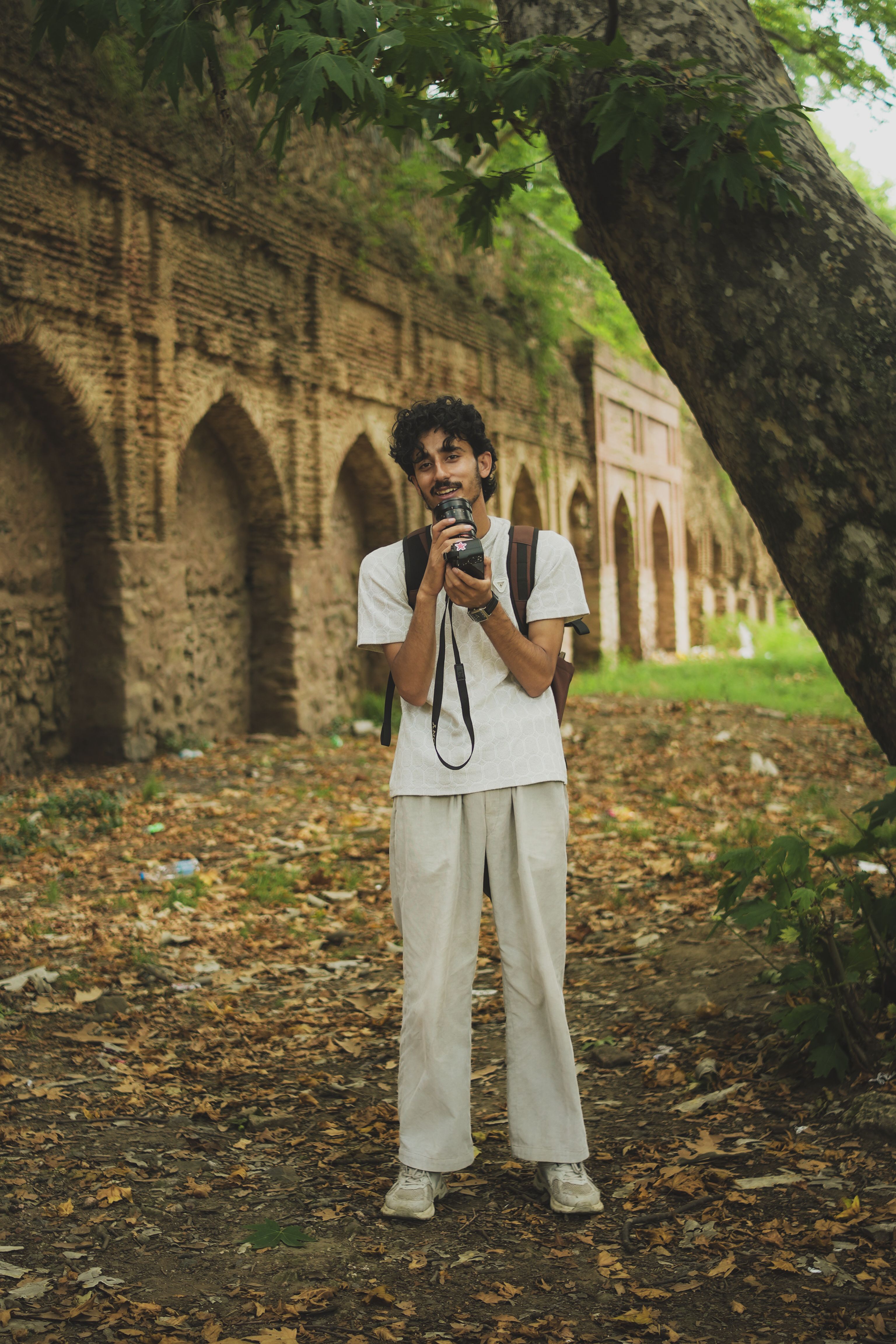 A young photographer holding a camera in a historic park surrounded by arches and trees.