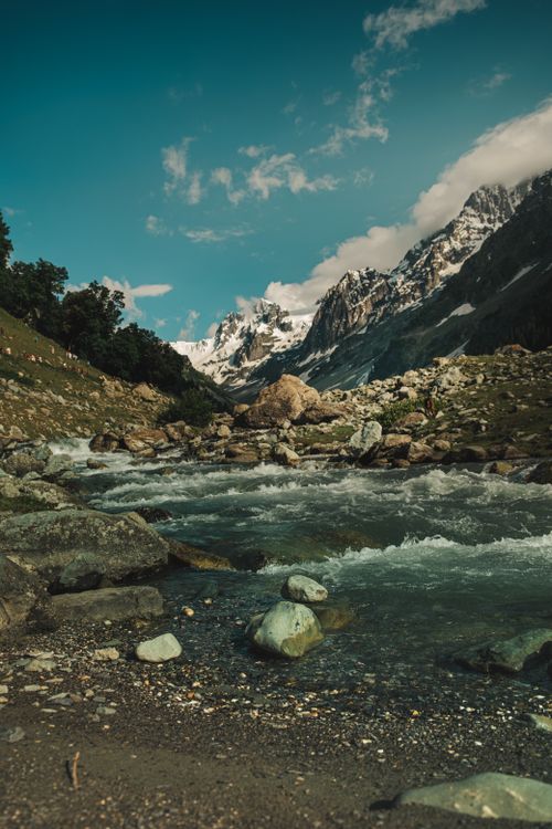 Free A serene mountain stream flowing through a rocky valley with picturesque snow-capped peaks in the background. Stock Photo