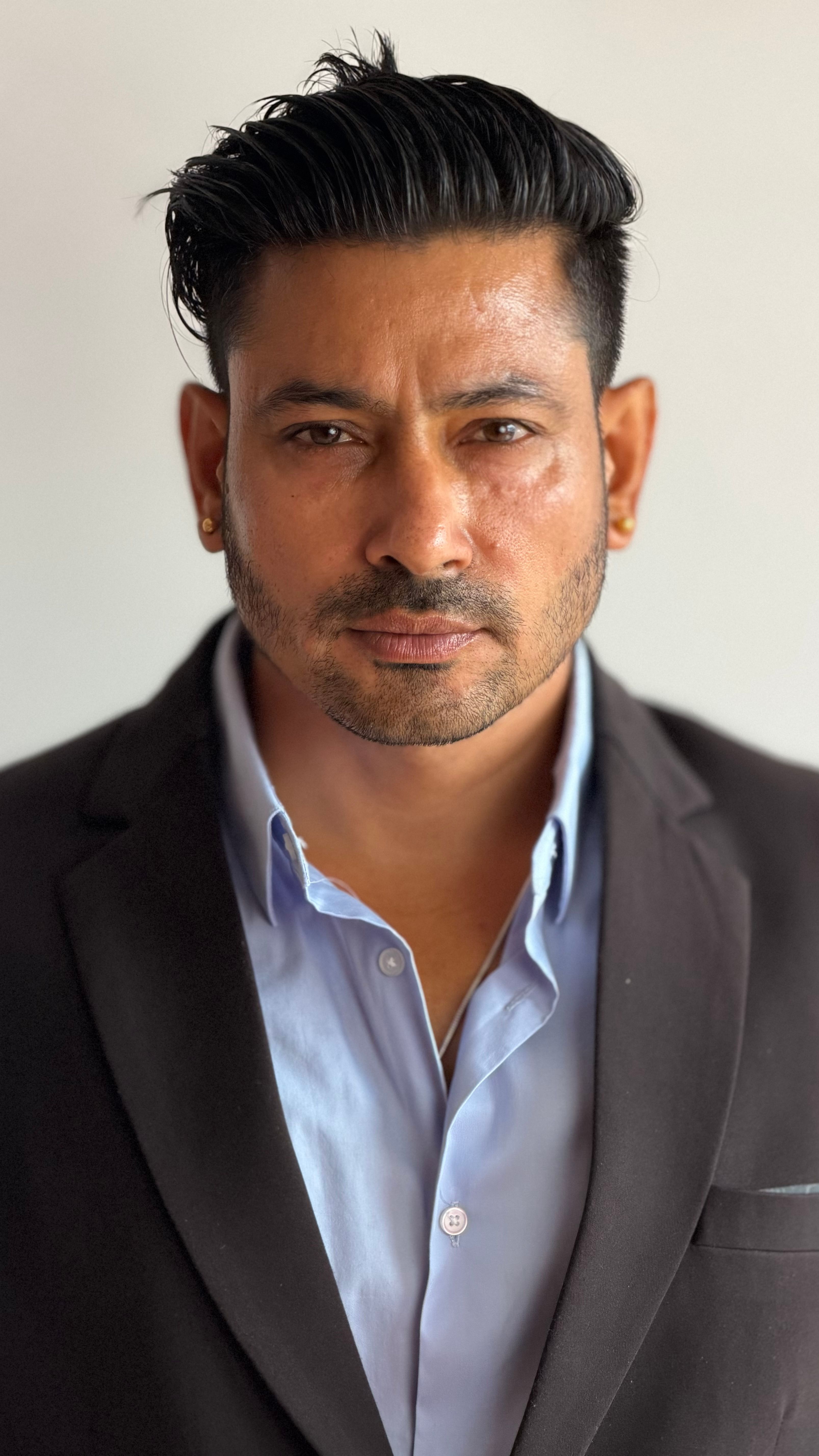A serious and confident man poses in a black suit and blue shirt against a plain background.