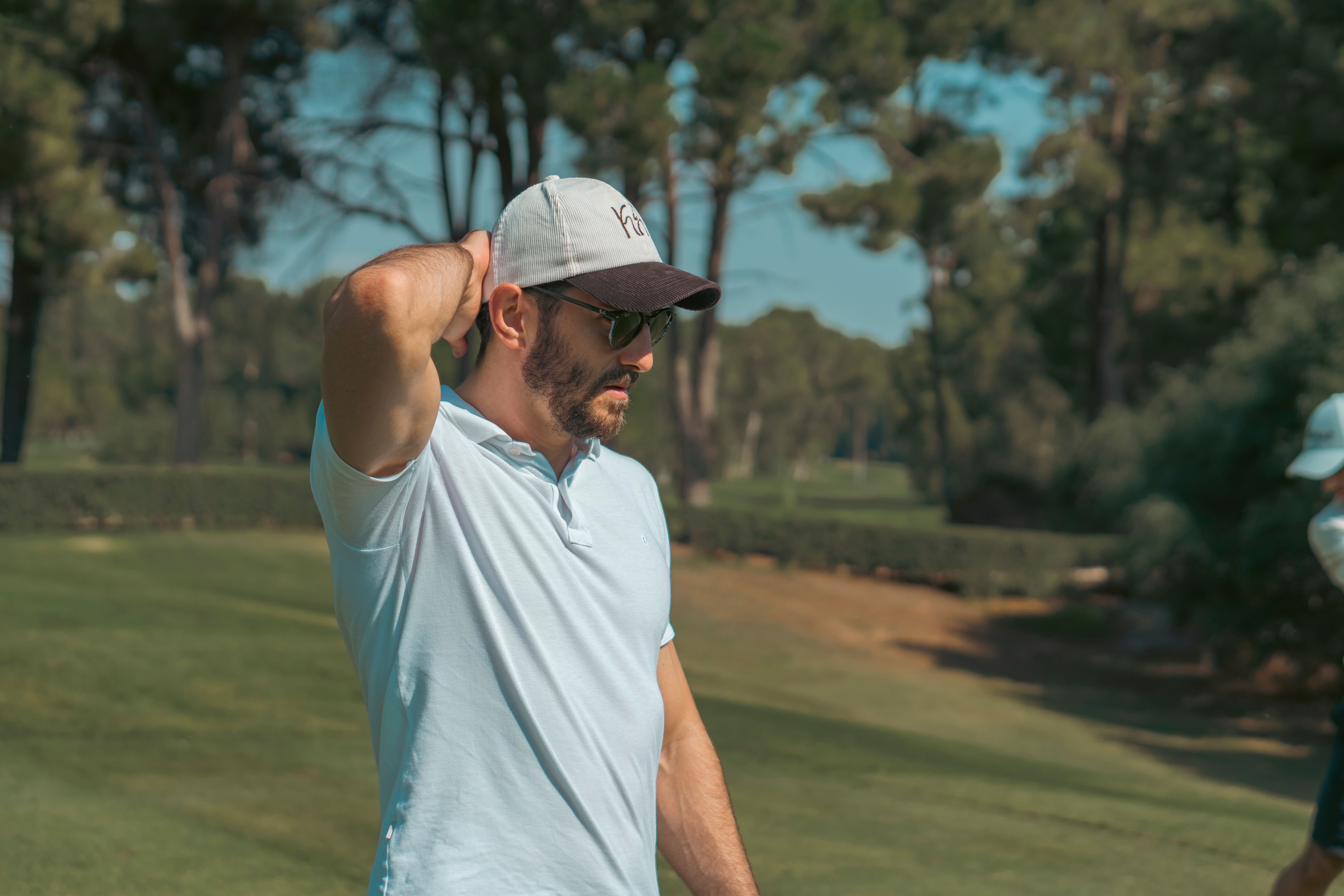 Man in casual attire wearing a cap stretches on a sunny golf course, surrounded by trees.