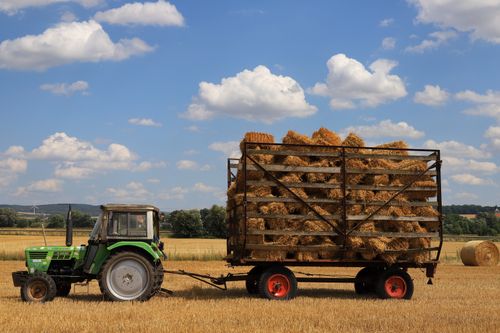 Free Green tractor towing a cart of hay bales in a sunny field under a blue sky with clouds. Stock Photo