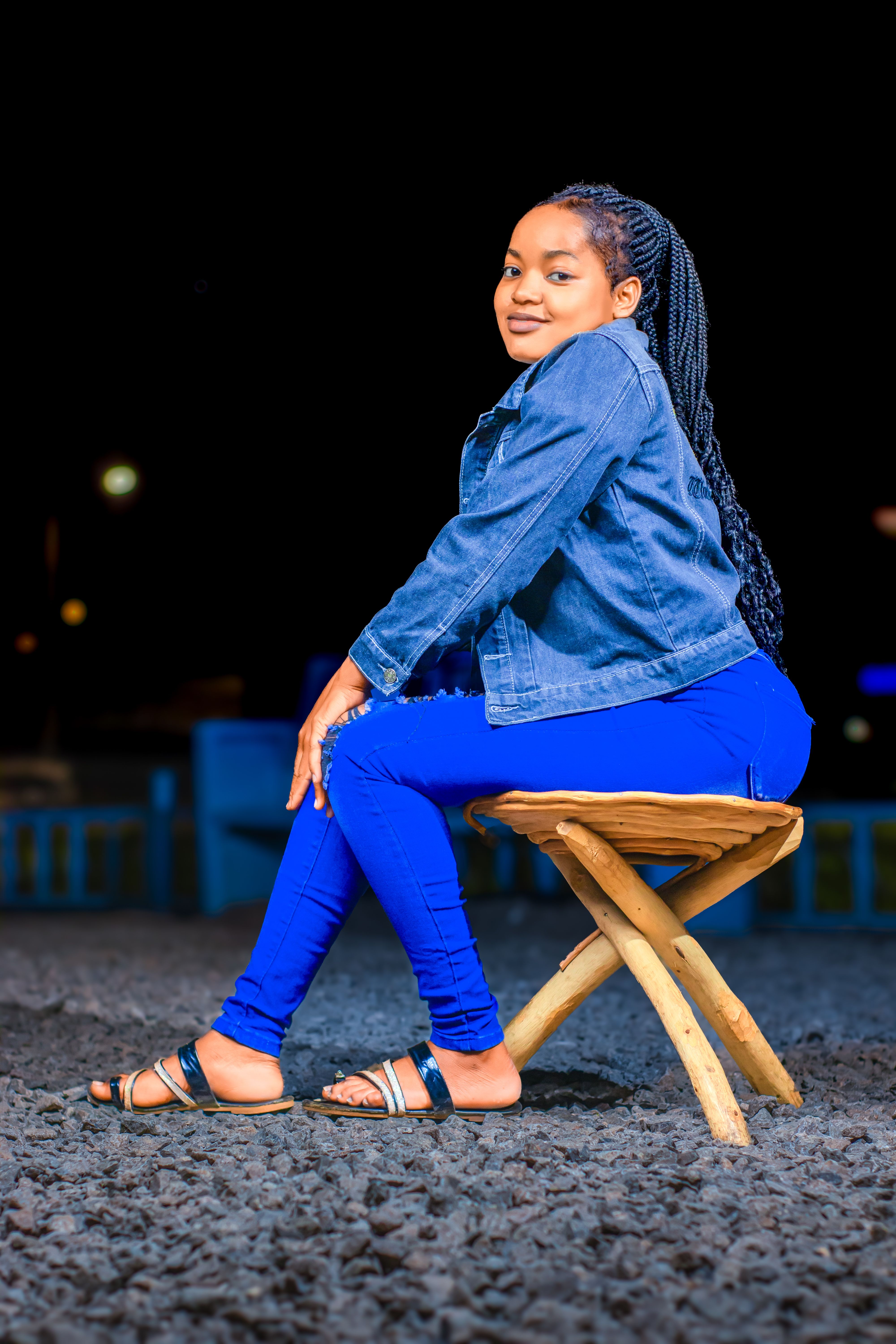 Stylish young woman in denim seated on a wooden stool outdoors at night.