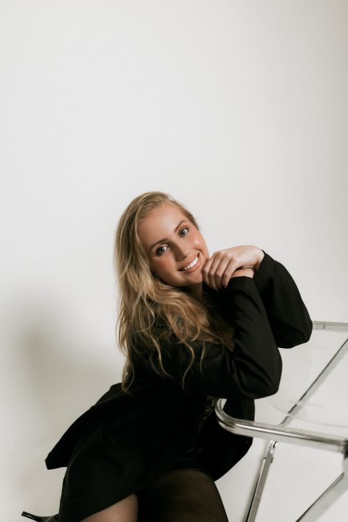 Free Portrait of a joyful young adult woman in stylish attire, leaning on a clear chair in a minimalist indoor setting. Stock Photo