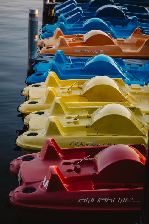 Free A row of vibrant paddle boats in various colors lined up on a calm body of water, ready for adventure. Stock Photo