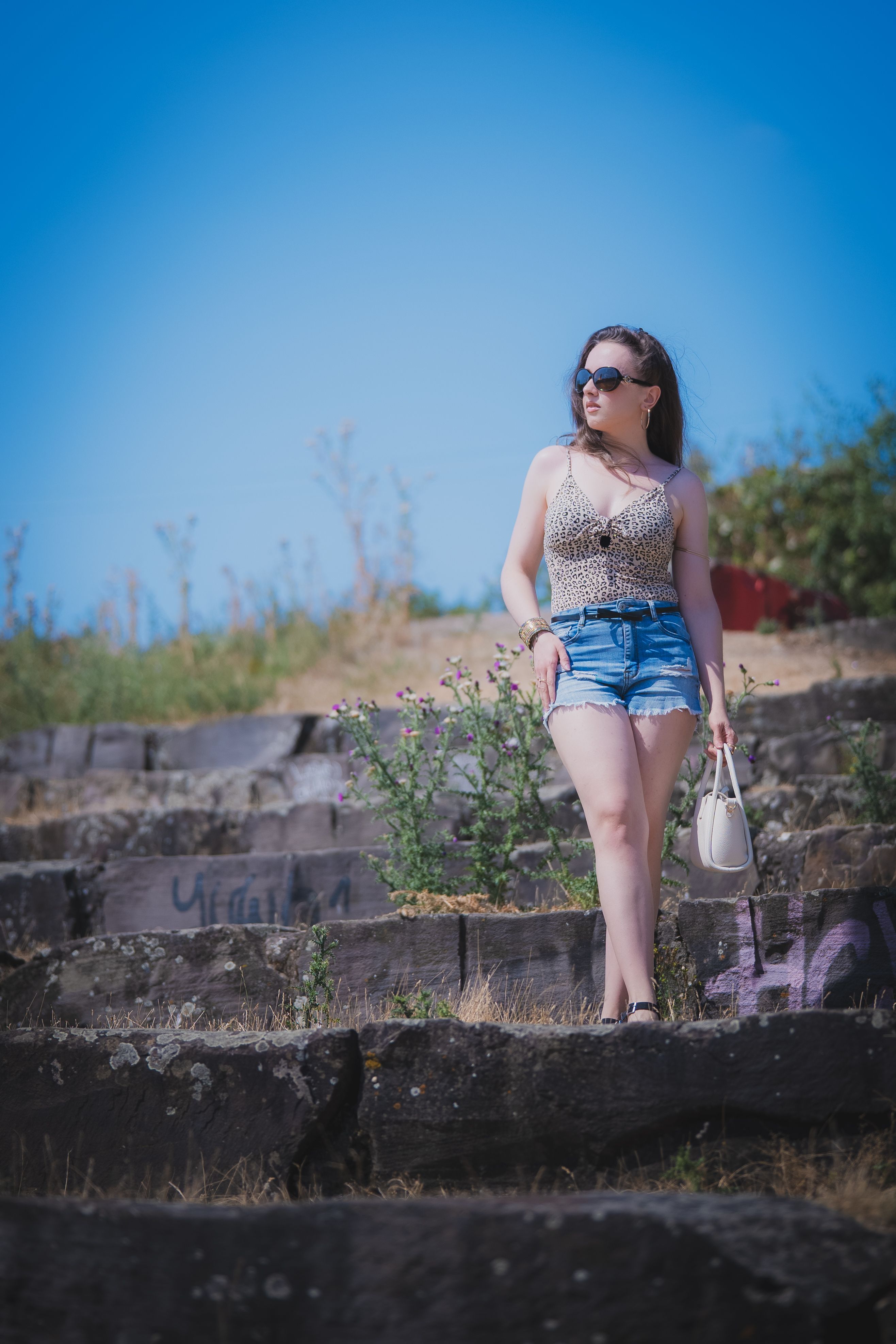 Uma jovem elegante, de óculos escuros, vestida com roupas de verão, está em pé com confiança em degraus de pedra ao ar livre.