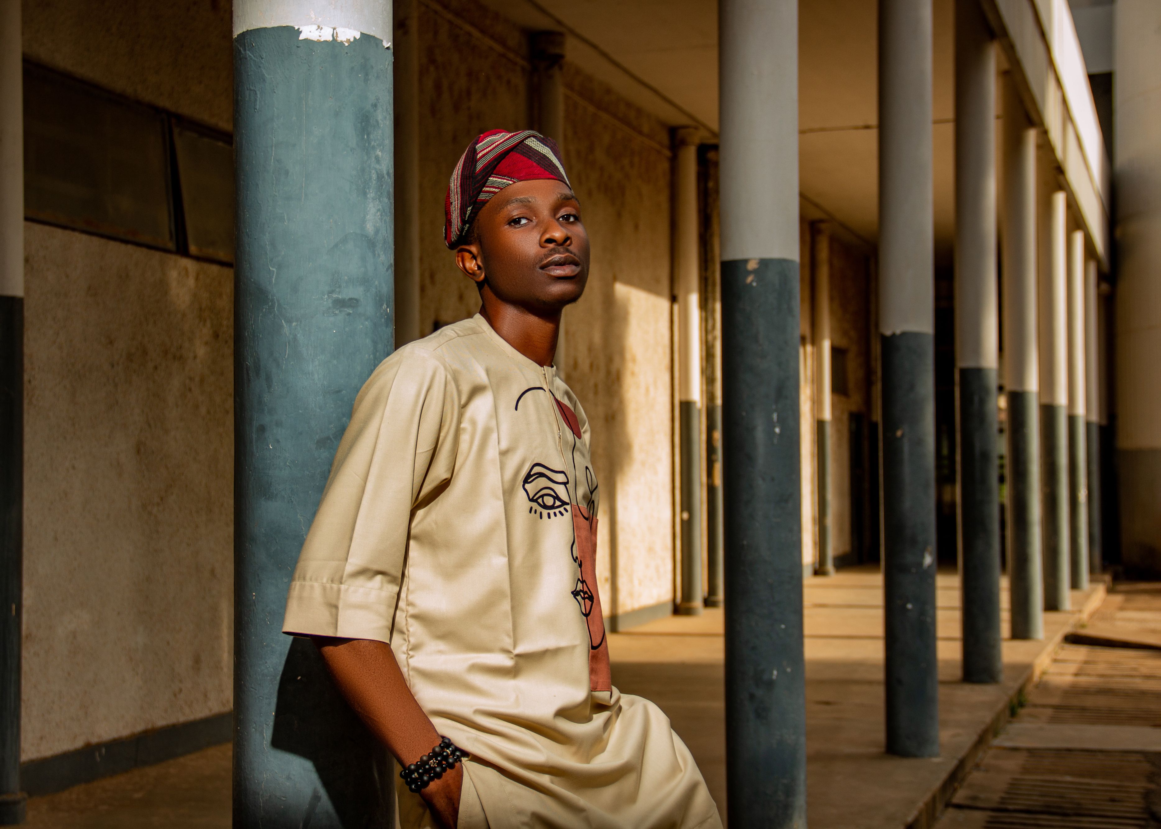 Portrait of a young man wearing traditional African clothing and headwear, leaning against a column in an outdoor corridor.
