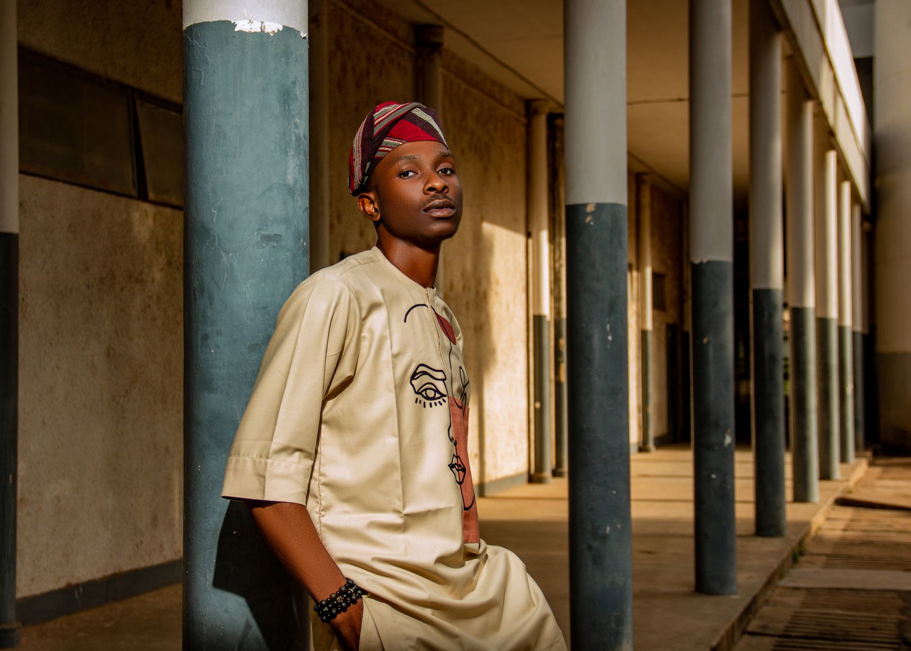 Portrait of a young man wearing traditional African clothing and headwear, leaning against a column in an outdoor corridor.