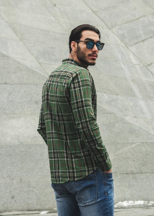 Man in Green and Black Plaid Shirt and Blue Denim Jeans Standing Near A Gray Concrete Wall