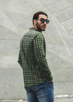 A stylish man wearing sunglasses and a plaid shirt poses casually outdoors against a modern concrete background.