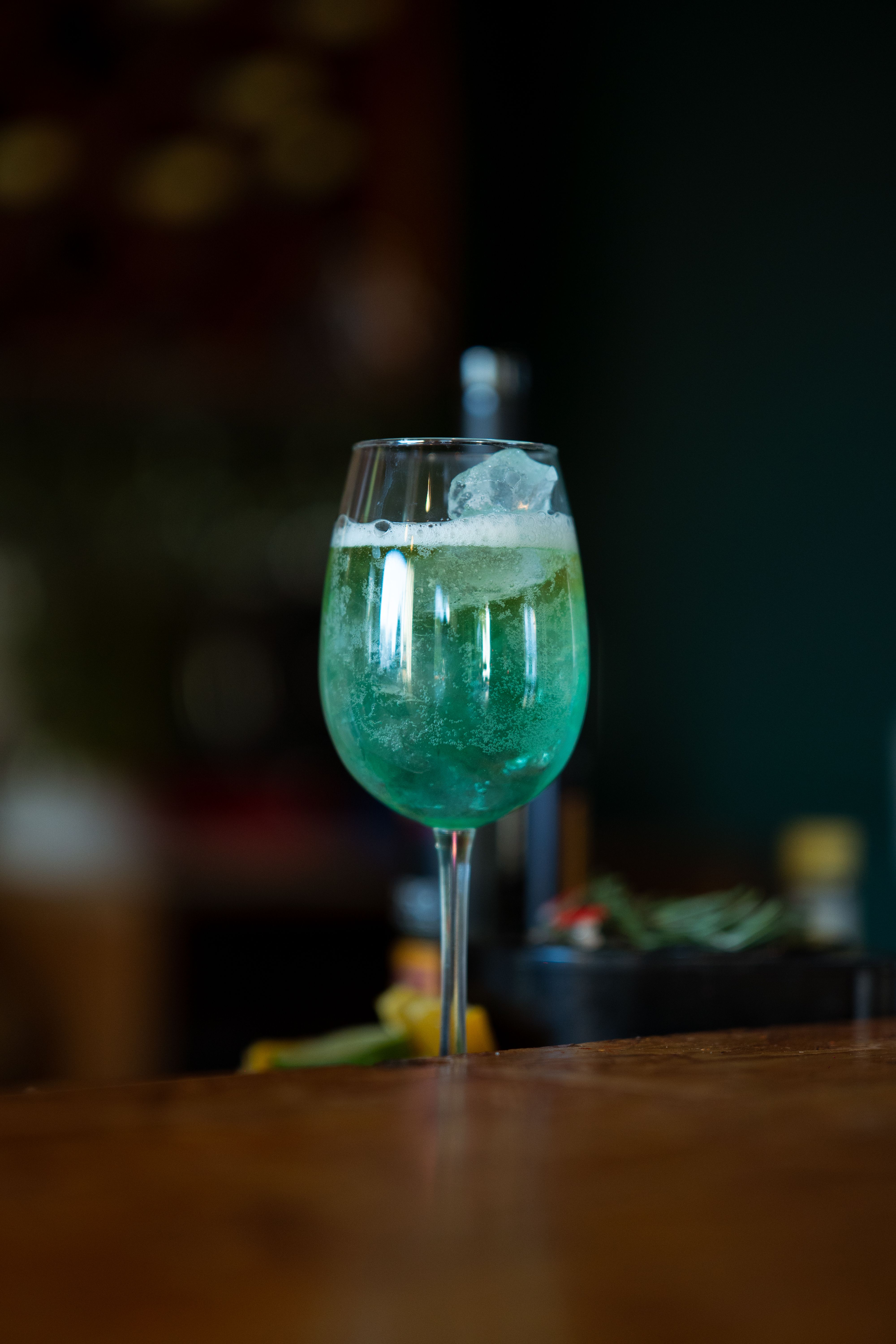 A vibrant green cocktail with ice served in a clear stemmed glass on a wooden bar.