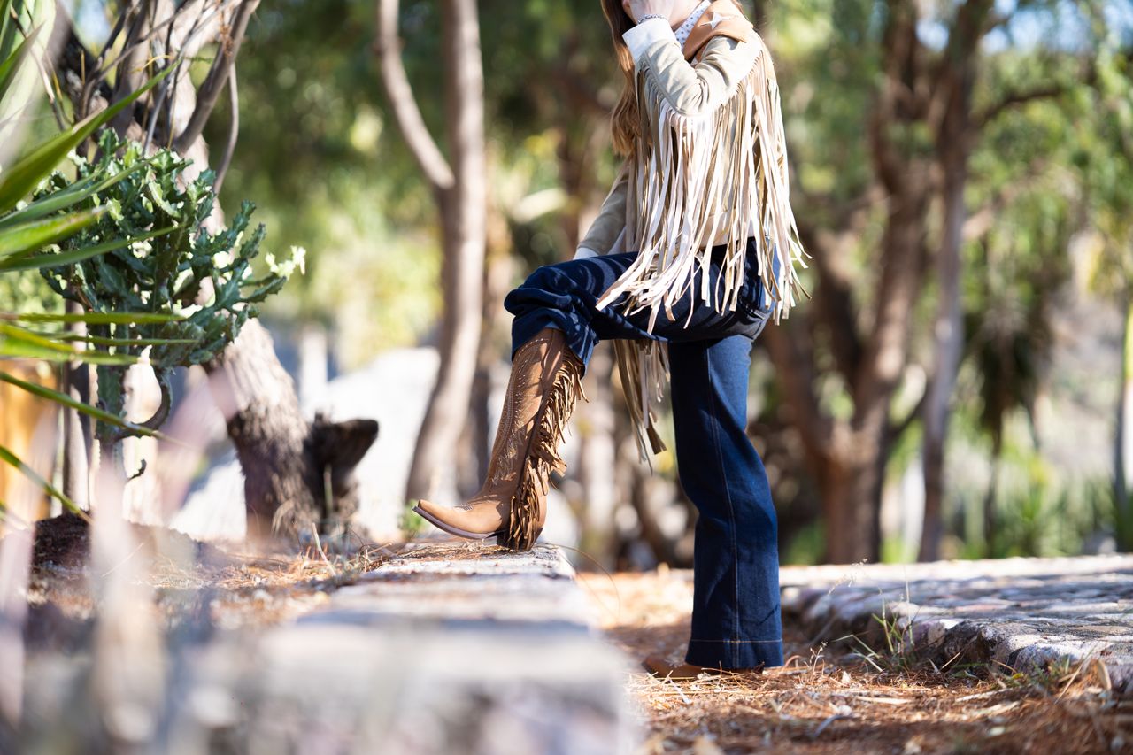 Woman in a cream fringe suede jacket with dark flared jeans and fringe cowboy boots among trees