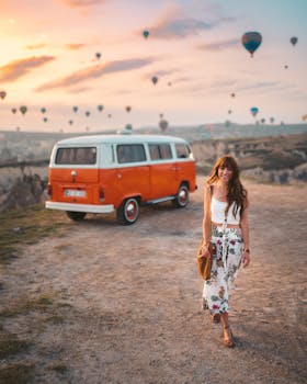A woman walking by an orange van during a vibrant sunset in Cappadocia with hot air balloons in the background.