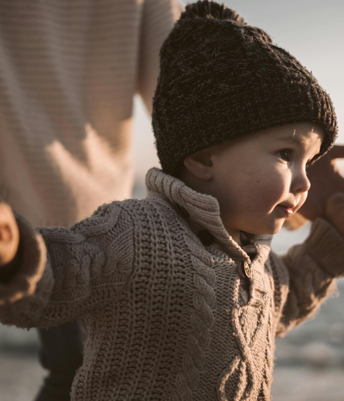 Close-up of a young child wearing a beige cable-knit sweater and a dark brown knitted hat