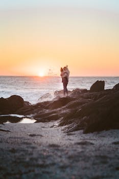 在岩石海滩上，一位母亲和孩子相拥着，面对充满活力的日落，这是一个感人的时刻