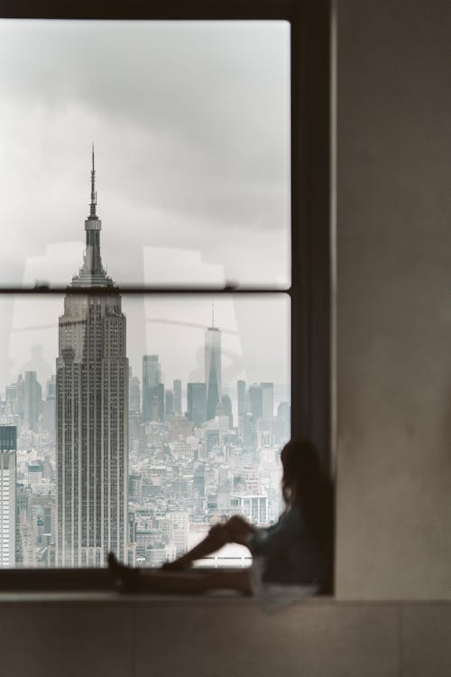 Free Woman Sitting at Window Sill Looking Out the Window at New York City Stock Photo