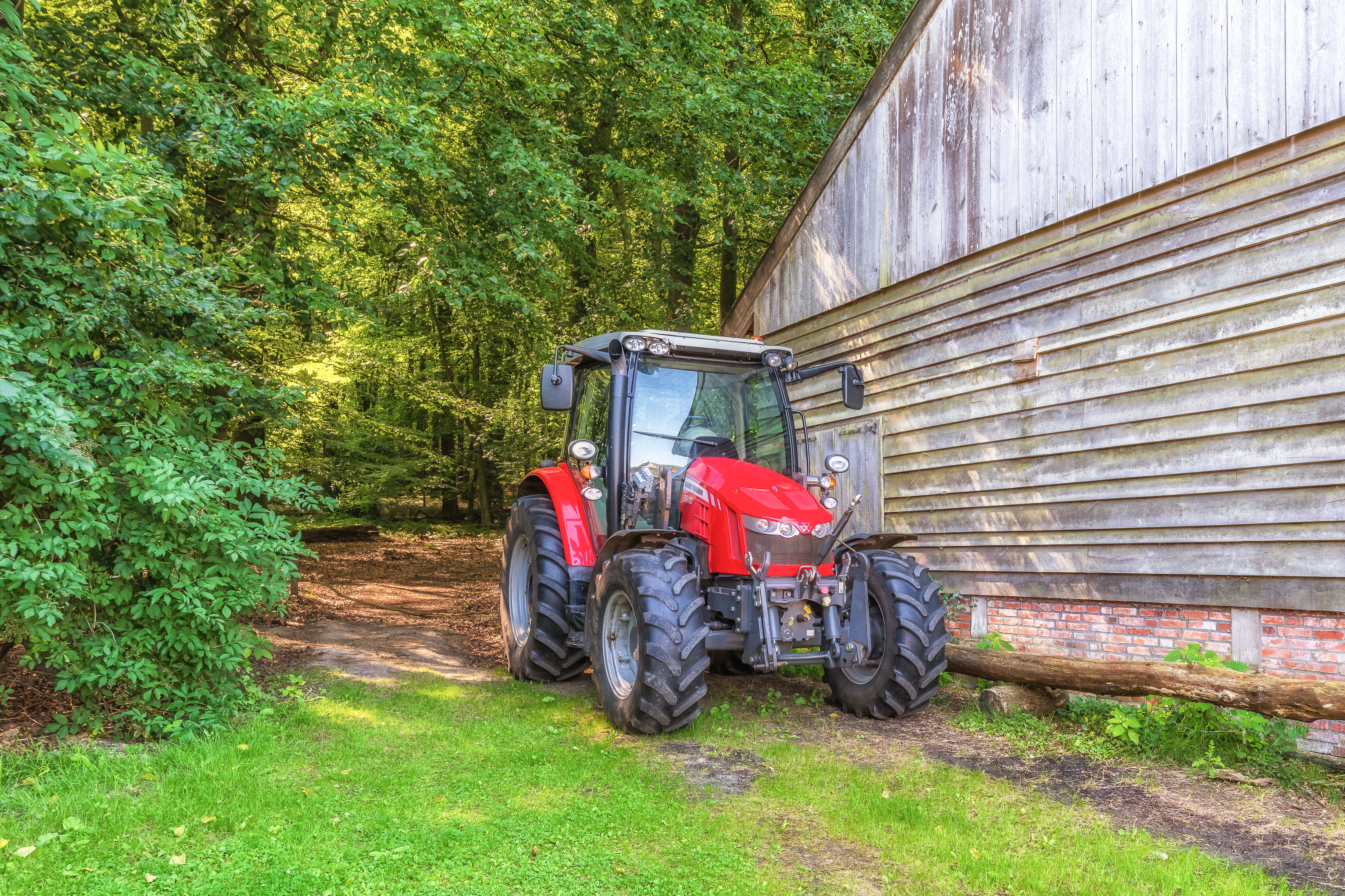 A vibrant red tractor parked beside a rustic wooden barn, surrounded by lush forest.