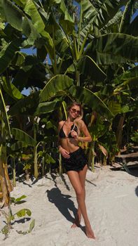 Adult woman in black swimwear posing on a tropical beach with lush greenery.