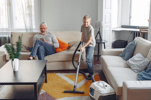 A boy vacuums the living room while an older man sits on the sofa using a phone.