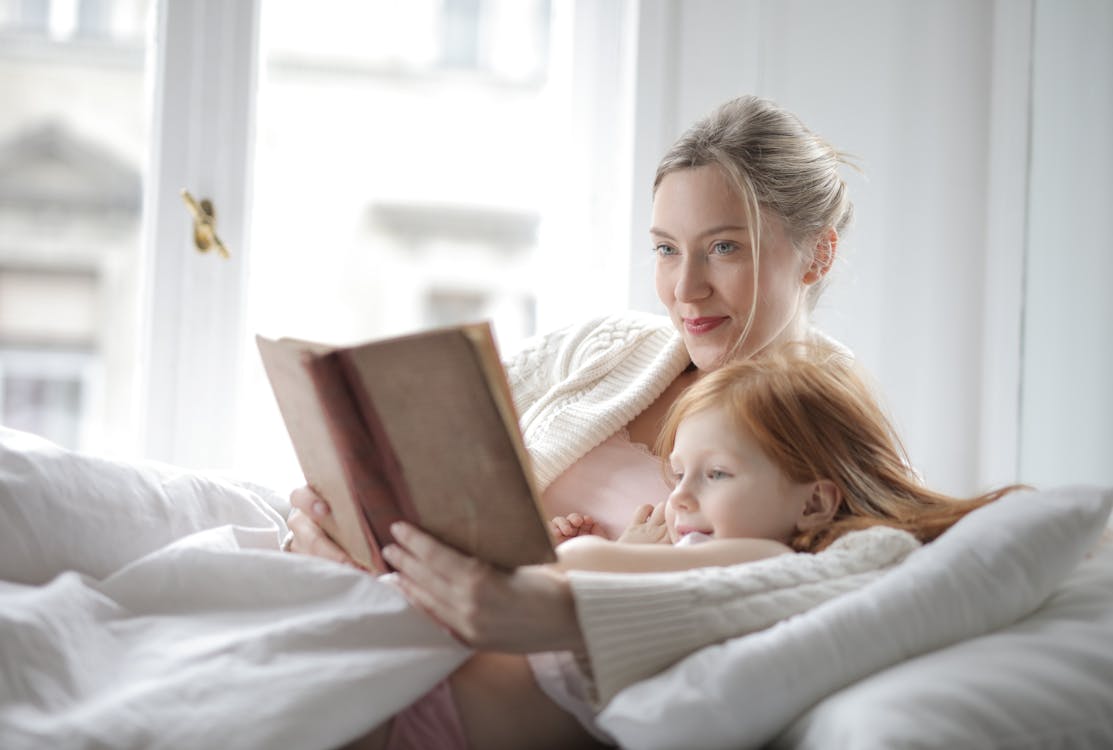 Woman in White Sweater Woman Holding Book Lying on Bed Beside Girl