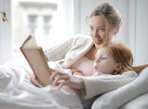 Free Woman in White Sweater Holding Book lying Beside Girl Stock Photo