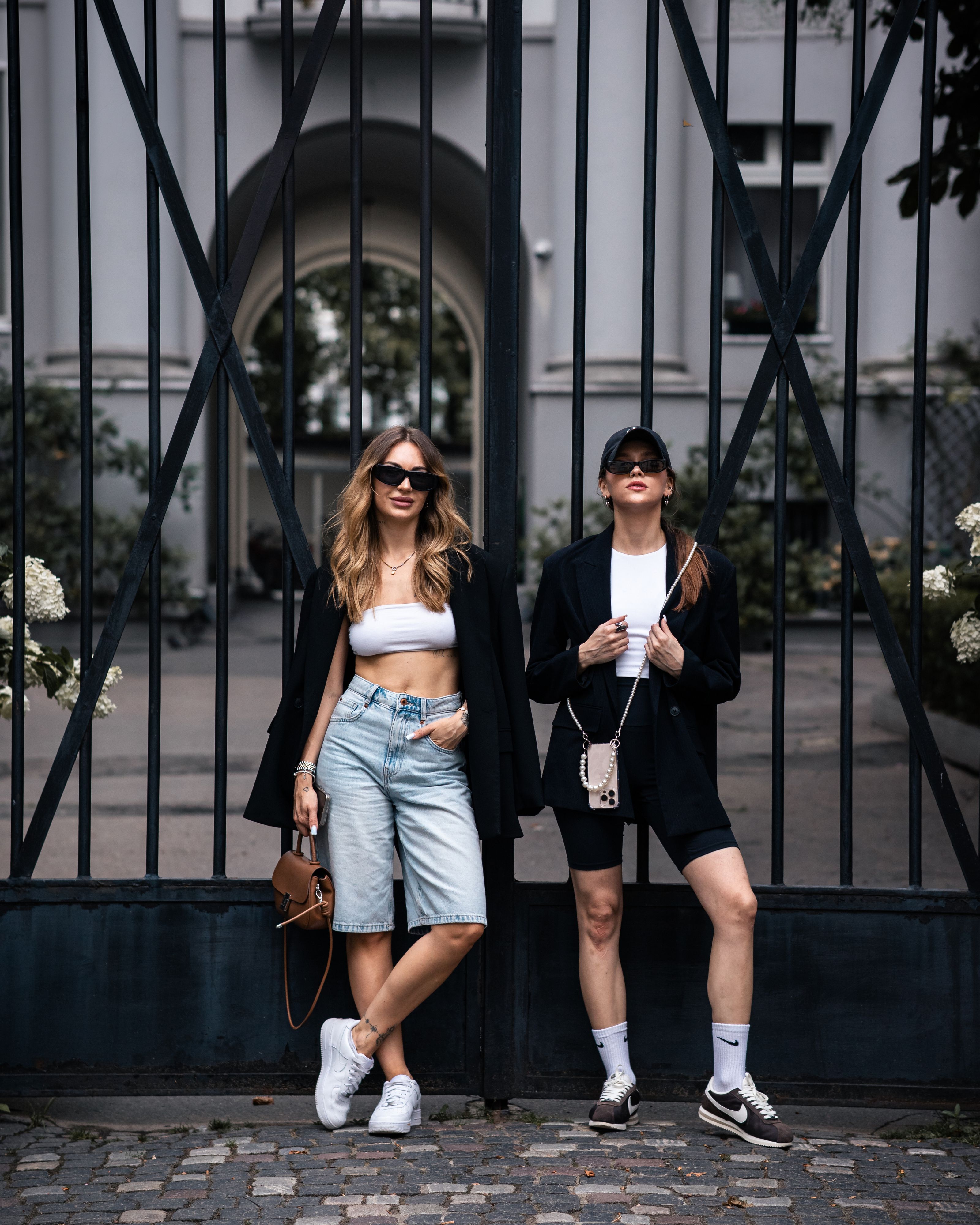 Duas mulheres estilosas posando com roupas de moda urbana em frente a um portão decorativo.