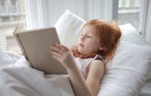 Free A young girl with red hair reading a book comfortably in bed, enjoying leisure time indoors. Stock Photo