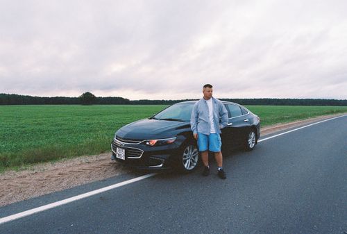 Free A man standing next to a black car on an empty rural road in Belarus near a green field under an overcast sky. Stock Photo
