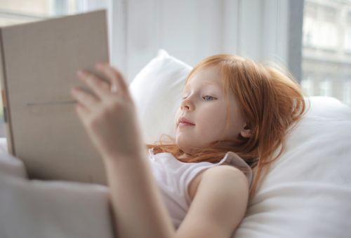 Free A young girl with red hair enjoys reading a book in a bright, cozy bedroom. Stock Photo