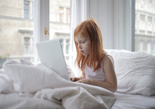 A young girl focused on a laptop while sitting on a comfortable bed in a bright bedroom.