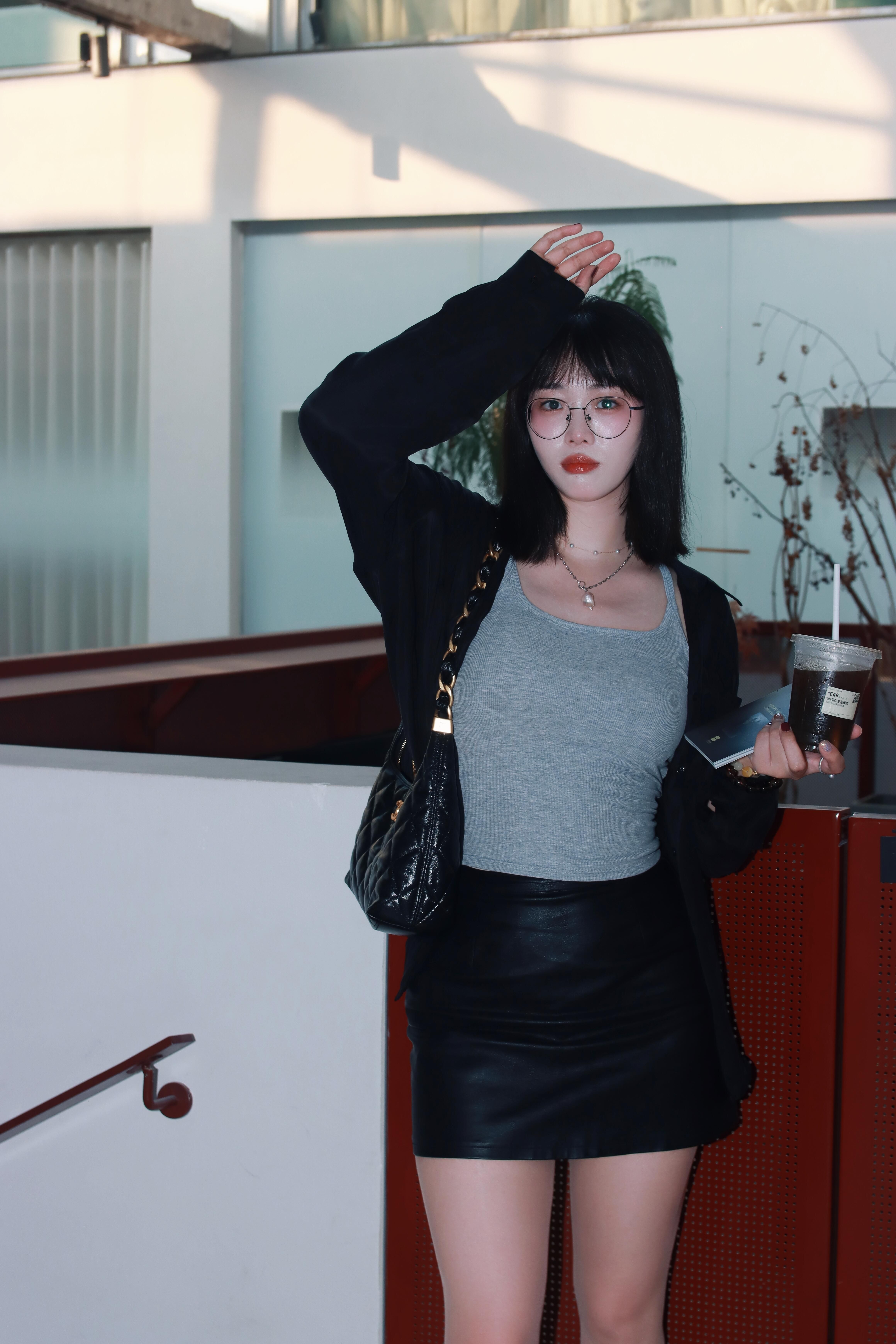 Stylish woman in urban setting, wearing a leather skirt and eyeglasses, holding coffee, indoors in Nanjing, China.