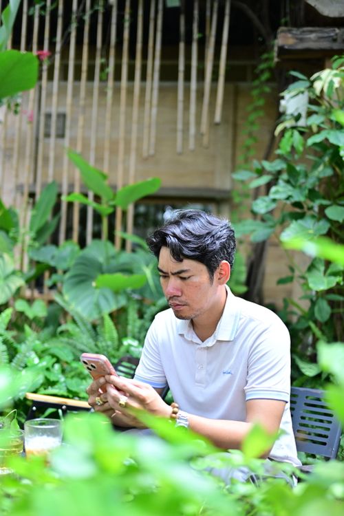 Free Young man focused on his smartphone, surrounded by vibrant tropical plants in an outdoor setting. Stock Photo