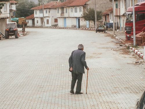 Free An elderly man with a walking cane strolls down a quiet cobblestone street in a rustic village. Stock Photo
