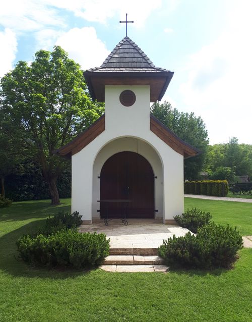 Free A serene small white chapel with a wooden door and cross, surrounded by lush greenery. Stock Photo