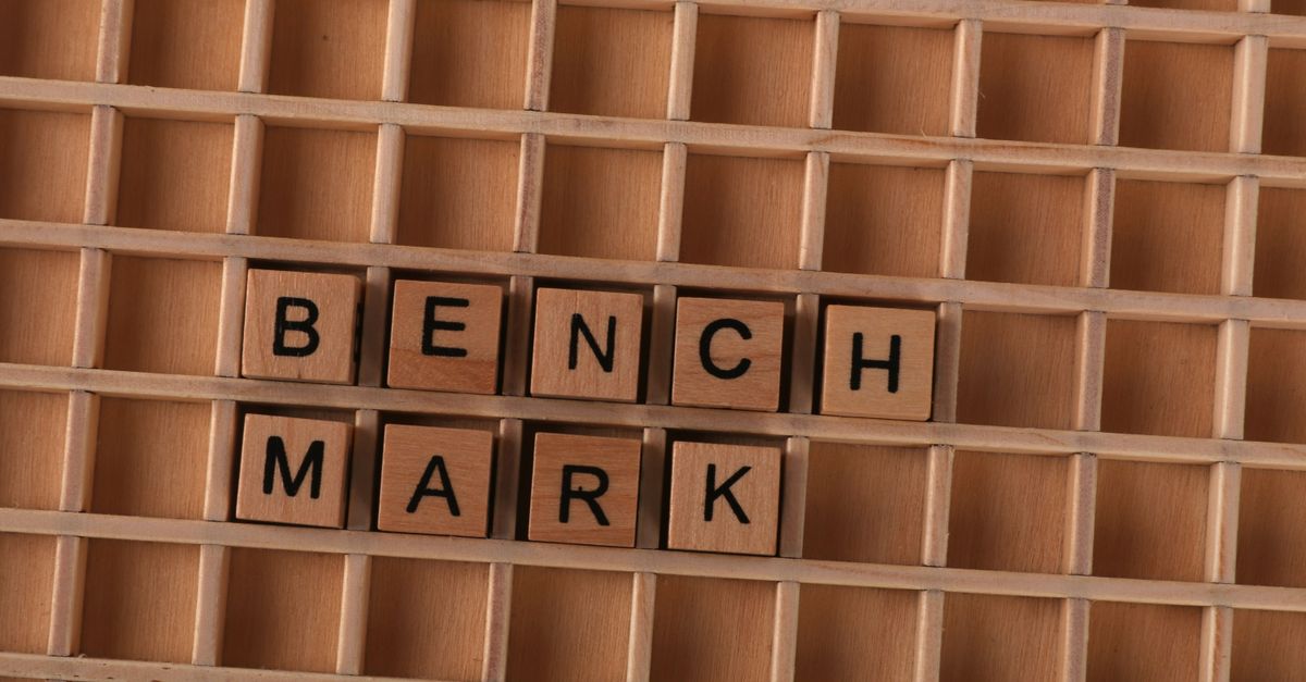 Wooden letter tiles spelling 'BENCHMARK' placed on a grid-like wooden background.