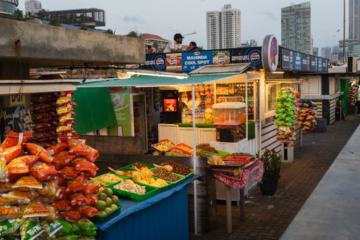 A vibrant street food stall in Colombo, Sri Lanka, offering snacks and fresh produce under city skyscrapers at dusk.