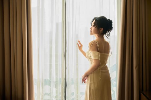 Elegant woman in a stylish dress looking through curtains, creating a serene and classy atmosphere.