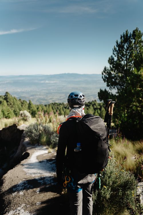 Free A hiker with gear stands on a scenic mountain trail overlooking a valley under a clear sky. Stock Photo