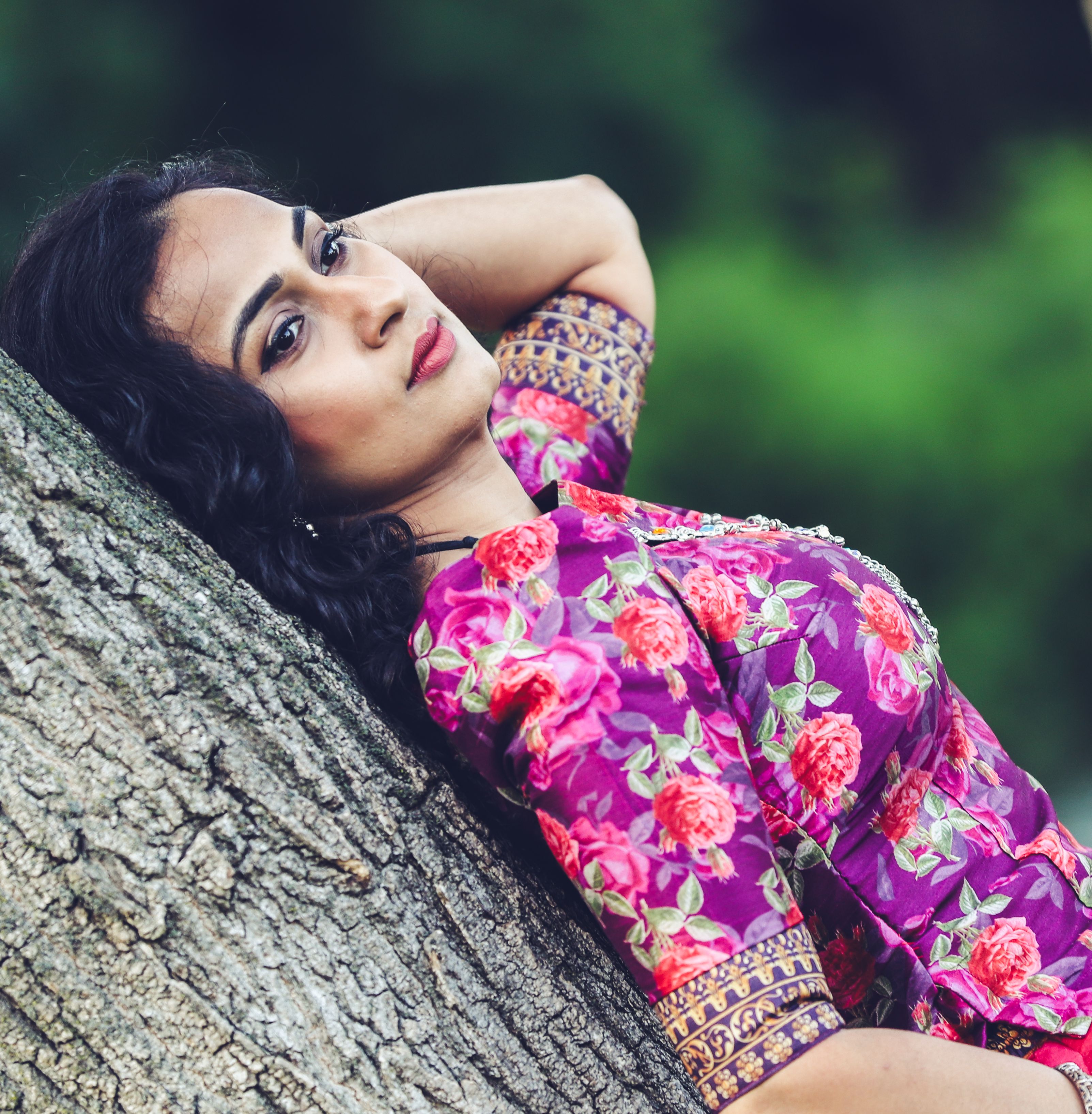 A woman in a floral dress leans gracefully against a tree trunk outdoors.