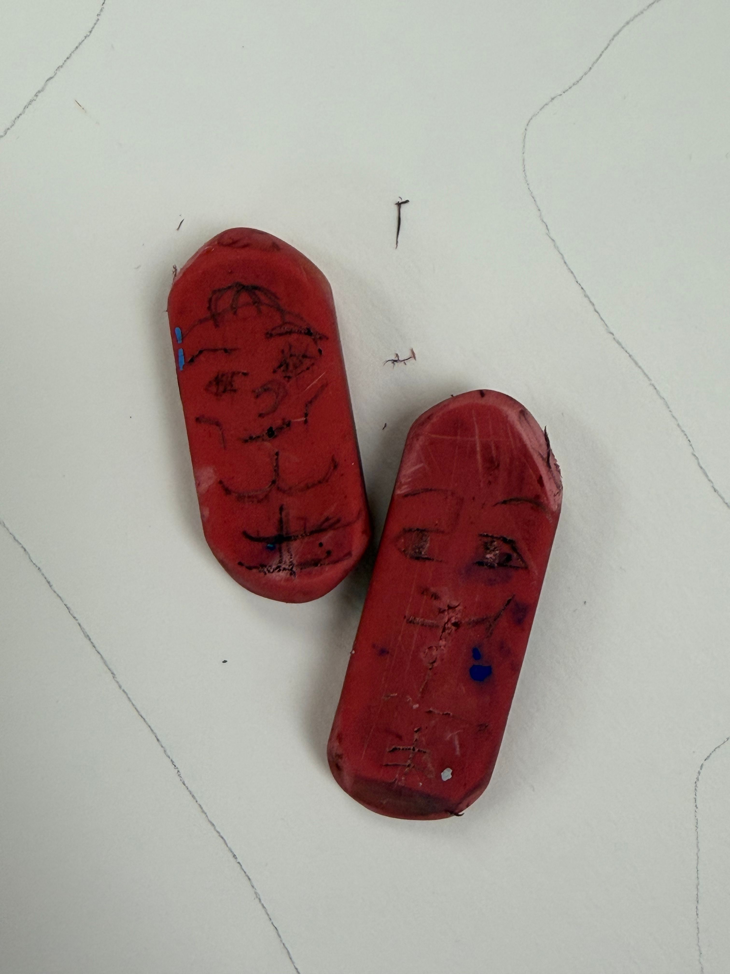 Two red erasers with artistic face doodles on a paper surface.