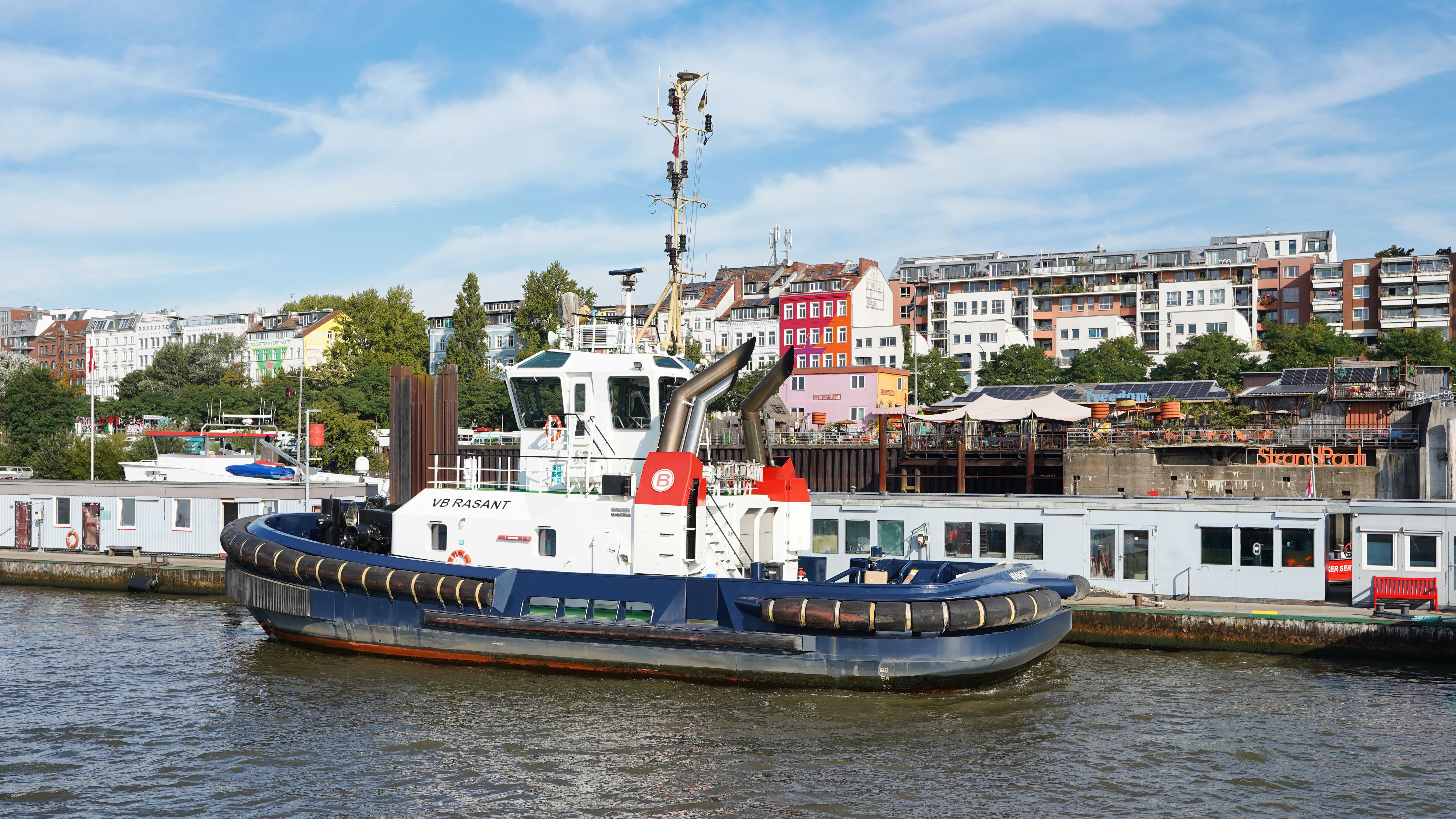 Tugboat VB Rasant docked in Hamburg harbor with colorful buildings in the background.