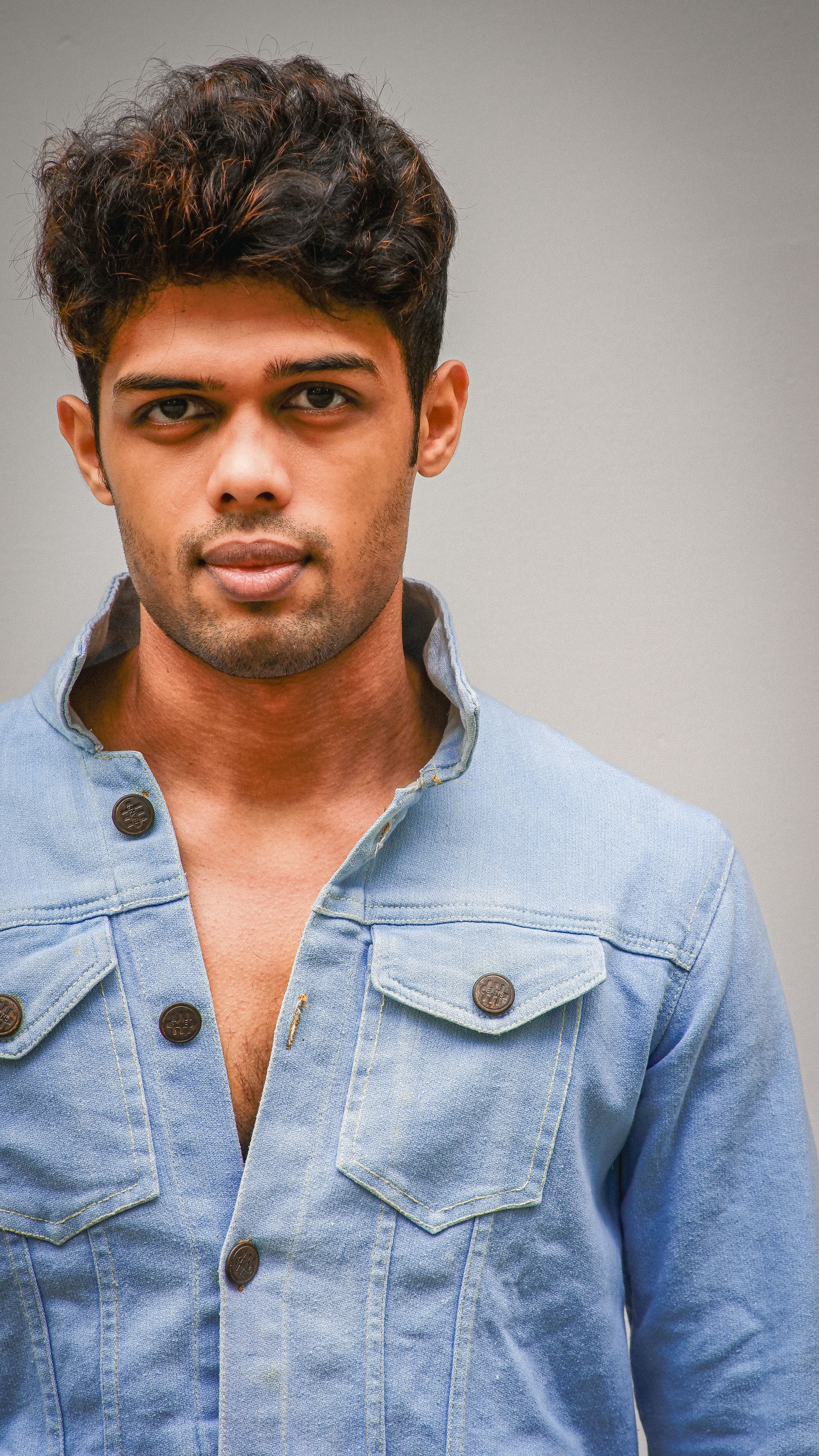 Close-up portrait of a young man wearing a denim jacket against a neutral outdoor background.