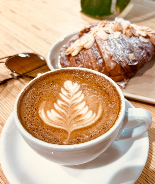 Free A delicious cup of latte with intricate foam art paired with an almond croissant, sunglasses on the wooden table. Stock Photo