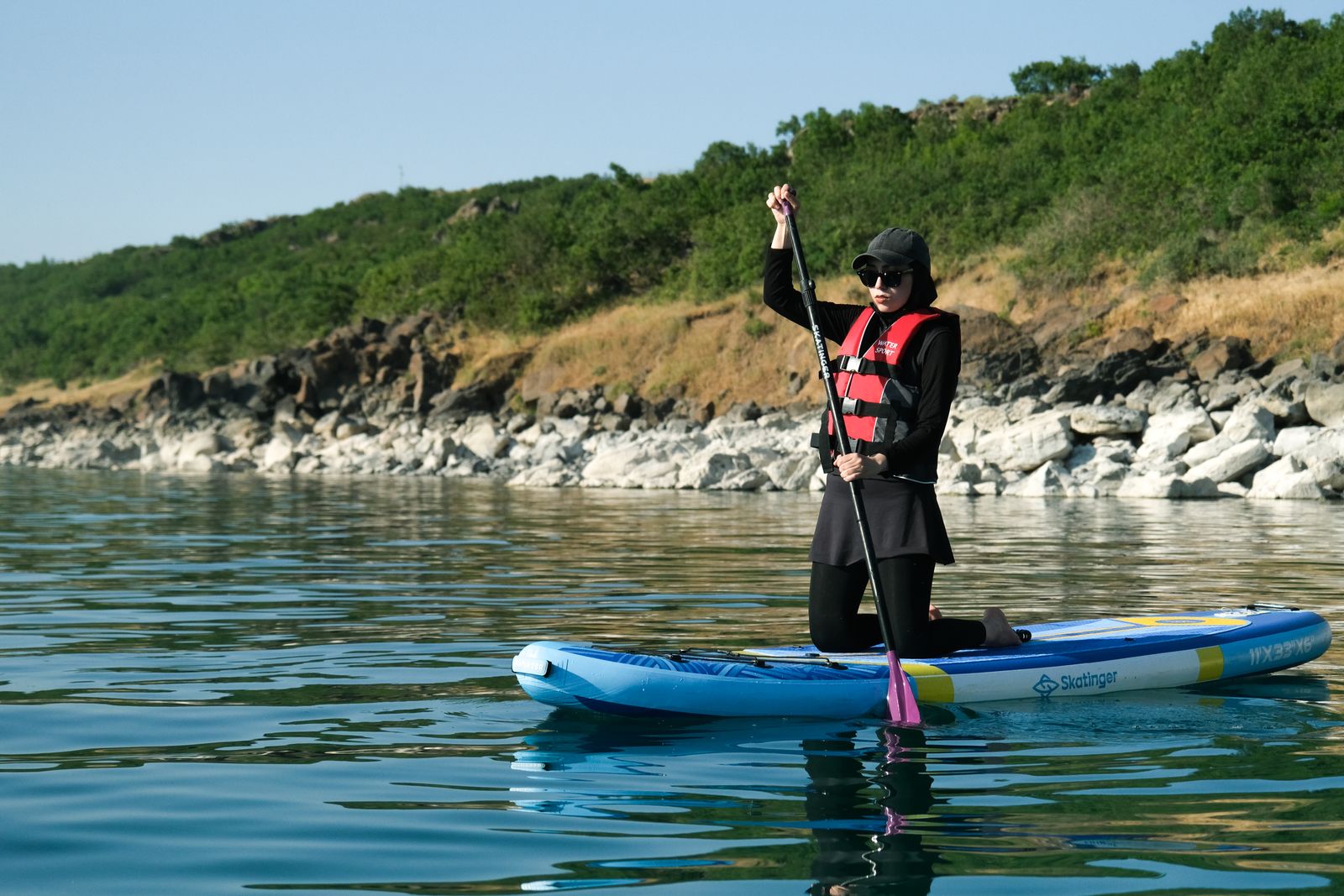 Woman wearing a life vest standing on an inflatable paddleboard and using a paddle on a calm lake
