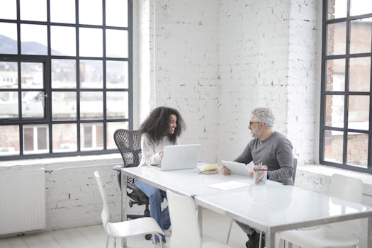 Two professionals collaborating in a bright, modern office space with laptops.