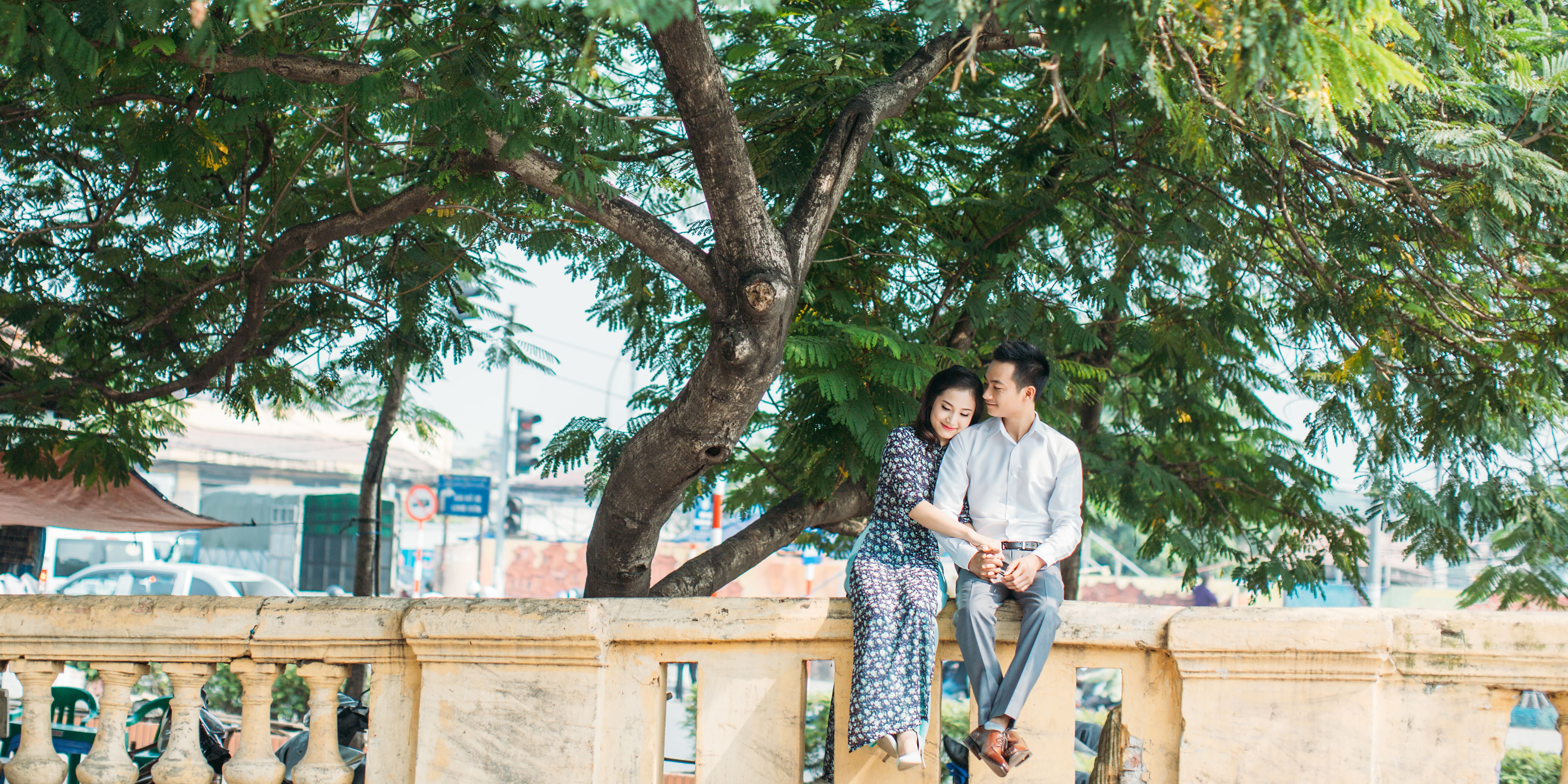 A couple shares a tender moment sitting on a stone bench under a lush green tree outdoors.