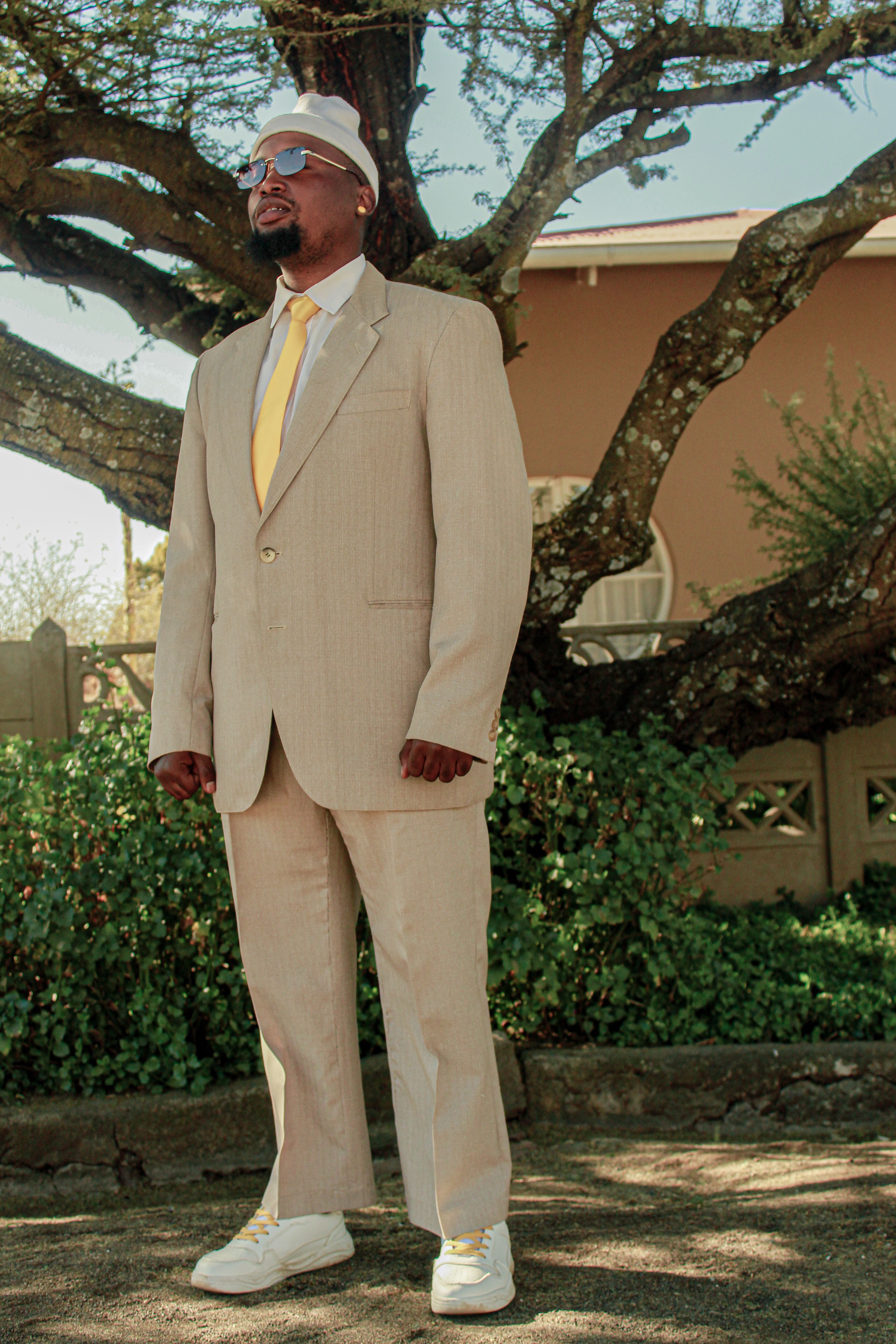 Man wearing a beige suit with yellow tie standing under a tree, exuding confidence.