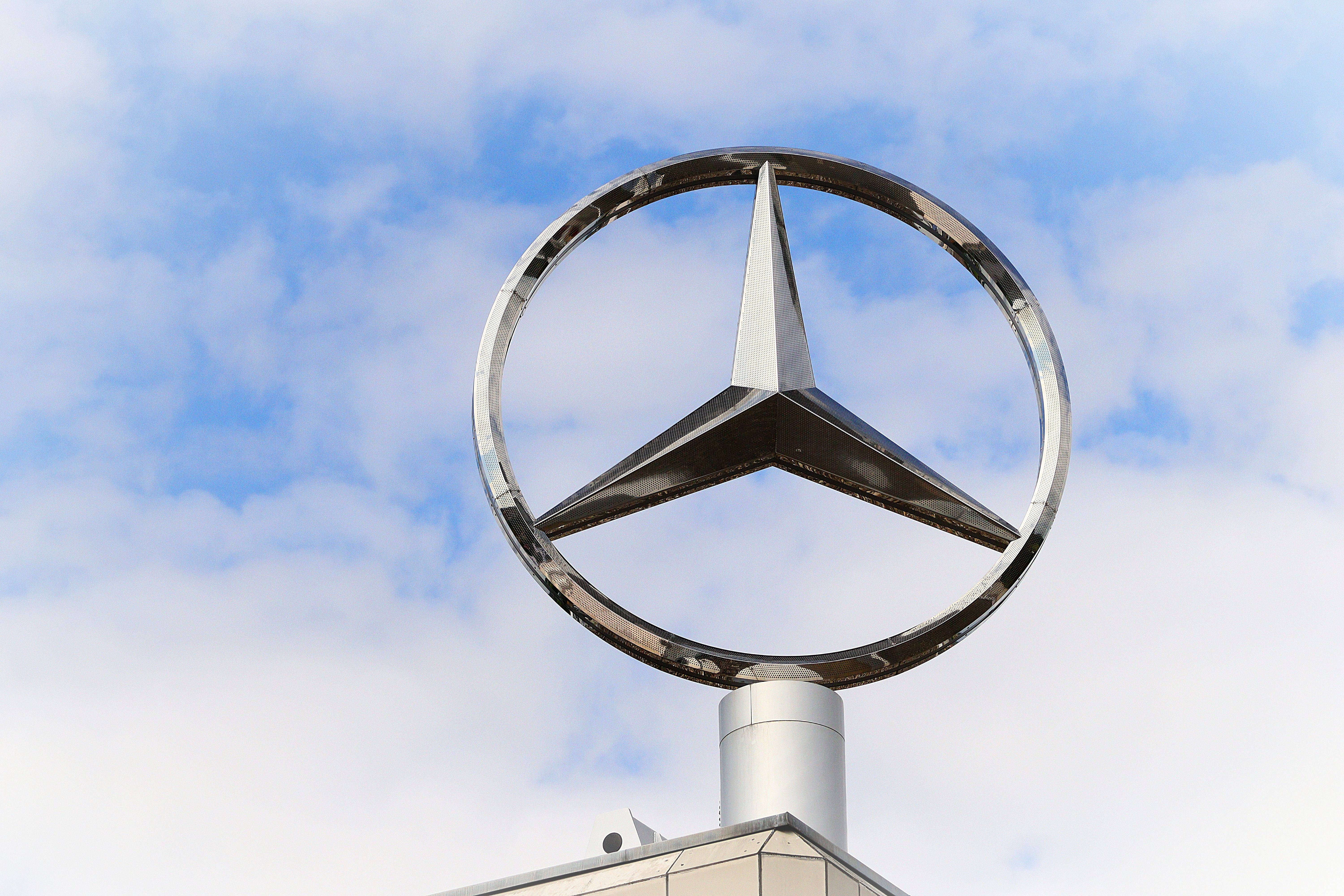 Stainless steel Mercedes-Benz logo against a blue sky with clouds.
