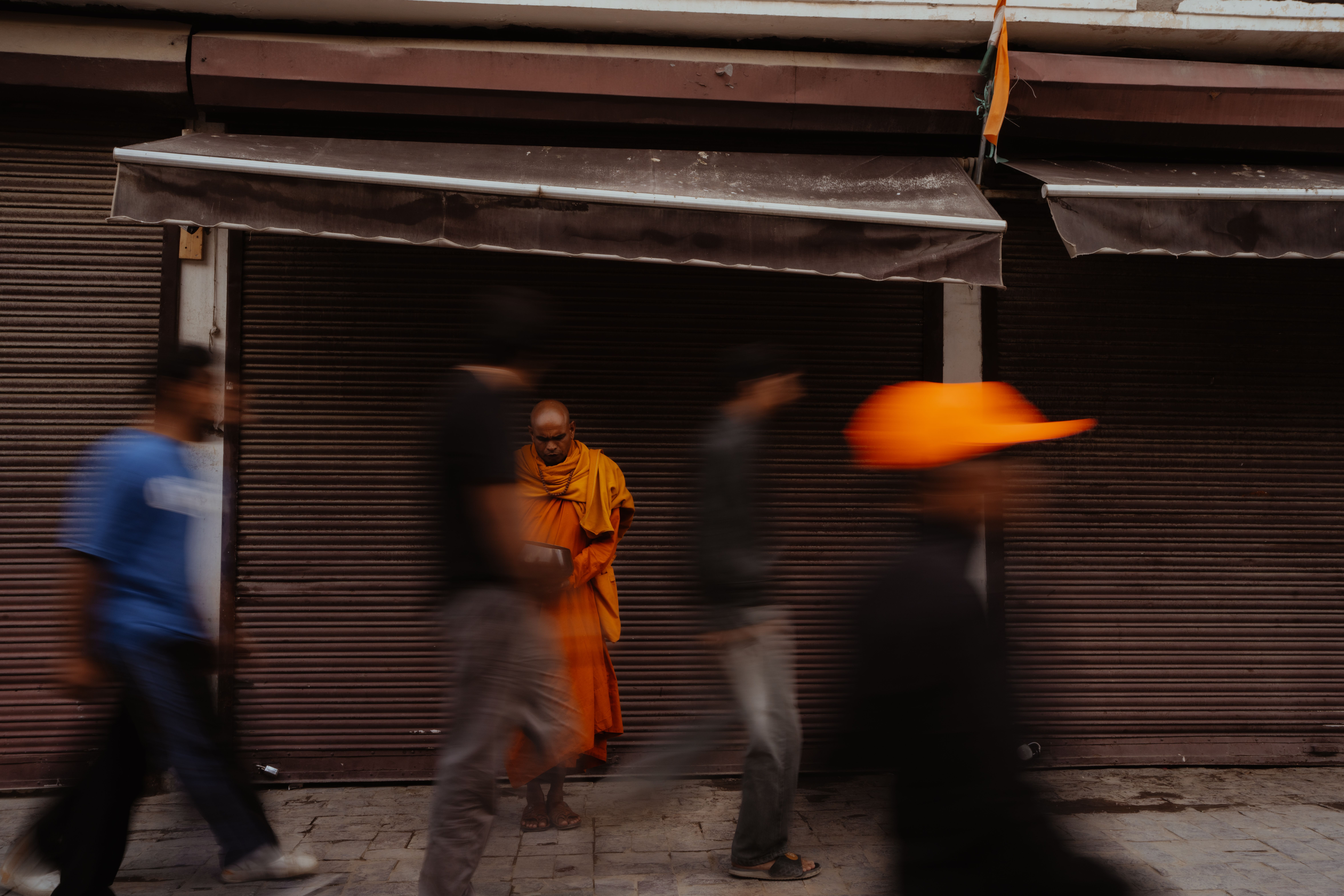 A monk in vibrant robes stands still as blurred pedestrians rush past in a busy street.